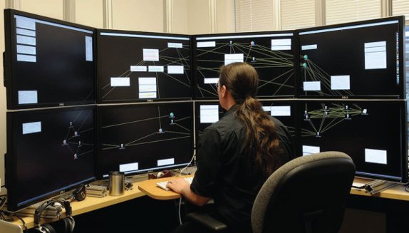 A person seated at a surround-screen display built from a grid of LCD monitors showing network graph visualizations.